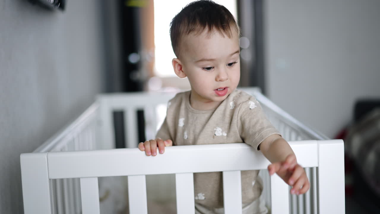 Adorable Caucasian kid stands inside his crib. Beautiful child stretches hands in front of him demanding something. Blurred backdrop.