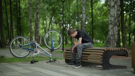 young boy wearing black top and grey pants sits with his head down, thoughtfully contemplating on park bench, his bicycle is turned upside down beside him, and a blurry background of trees