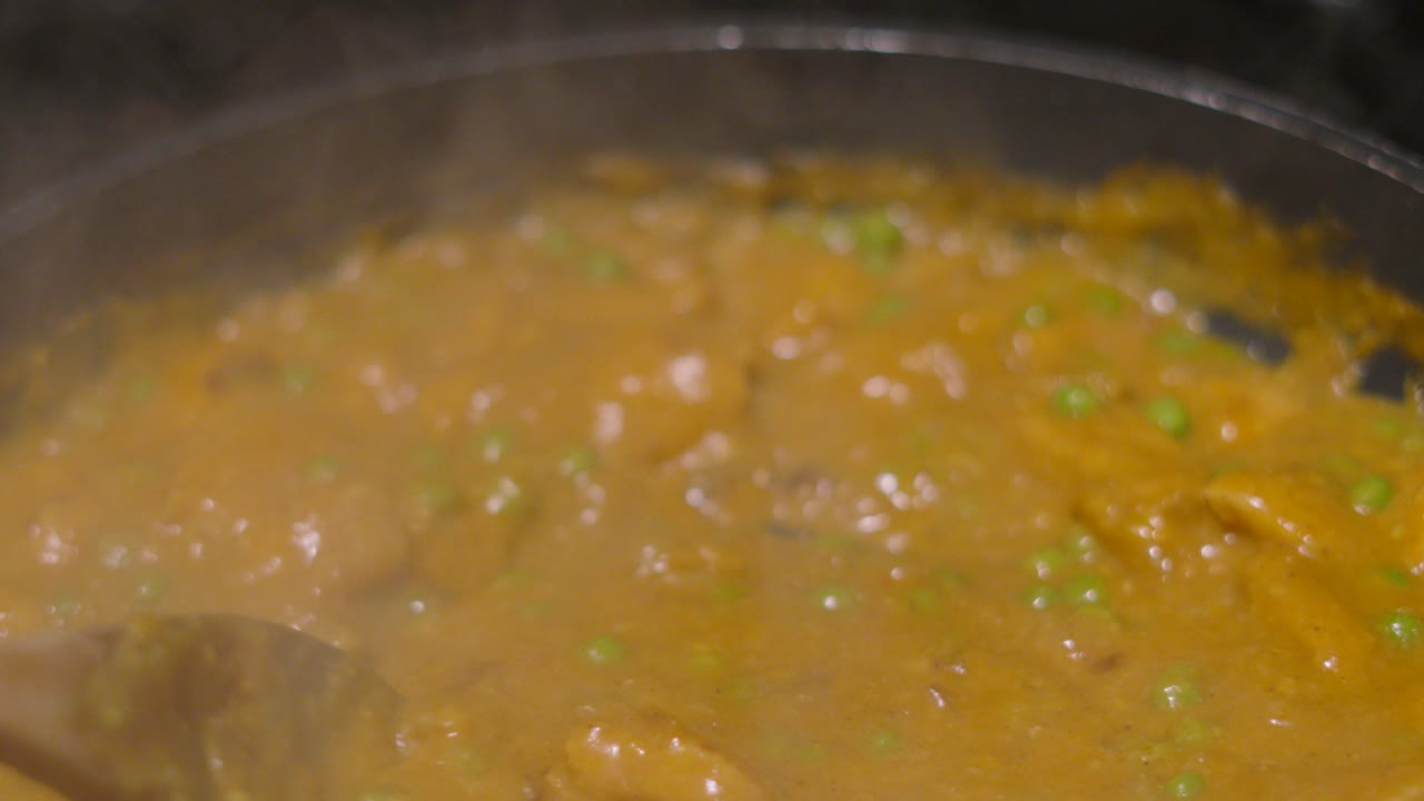 Nærbillede af en kok, der bruger en træske til at røre en cremet chicken korma-karry i en slip-let stegepande. Billede i fugleperspektiv, der viser damp og boblende sovs