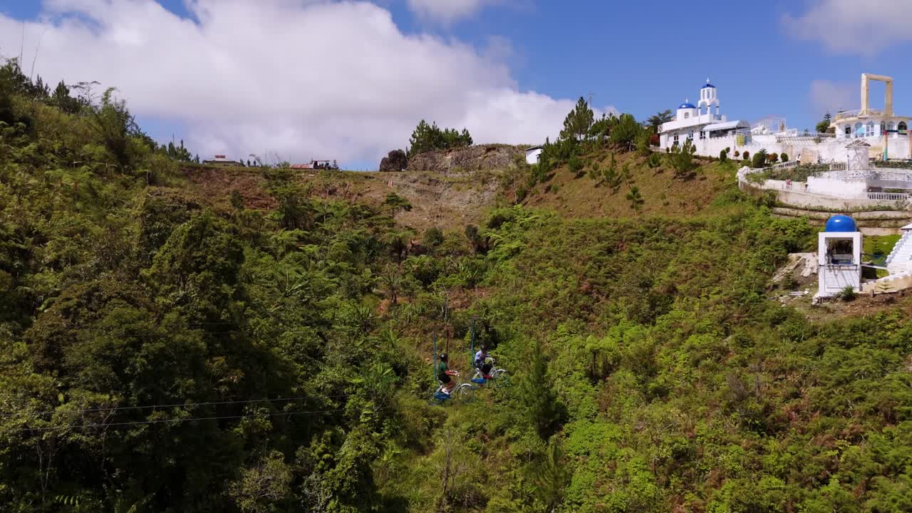 Drone footage of tourists riding sky bikes above lush green hills near a white hilltop landmark in the Philippines, showcasing adventure tourism, tropical scenery, and a bright outdoor landscape