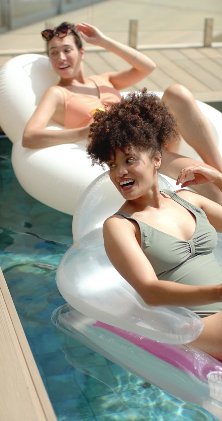 Vertical video: Two women lounging on lounge at pool in swimwear, holding cup, spotting off-frame