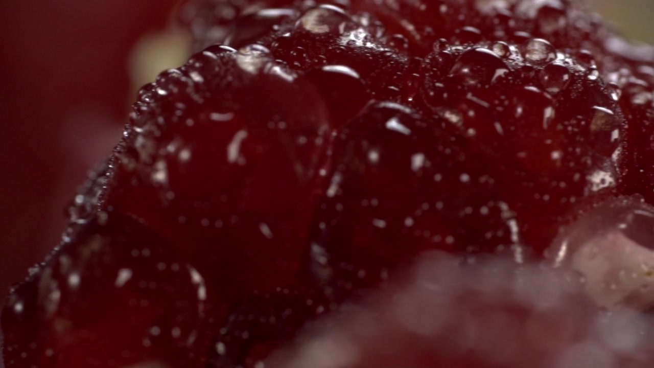 Primo piano estremo di un arillo di melograno fresco che mostra una superficie rossa lucida, polpa traslucida e goccioline di succo naturale con texture dettagliata e colore ricco.