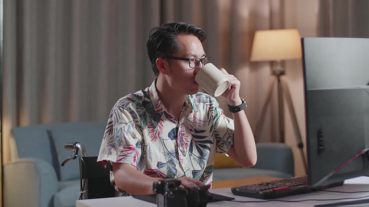 Uomo asiatico in sedia a rotelle che beve caffè mentre monta il video con un computer desktop accanto alla telecamera a casa