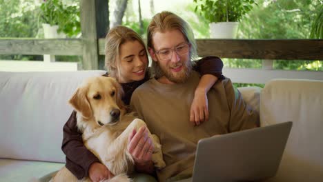 A-blond-girl-and-a-blond-guy-with-a-beard-in-glasses-are-looking-at-something-funny-on-a-laptop-and-laughing-next-to-them-their-dog-is-sitting.-Leisure-in-the-gazebo-in-nature