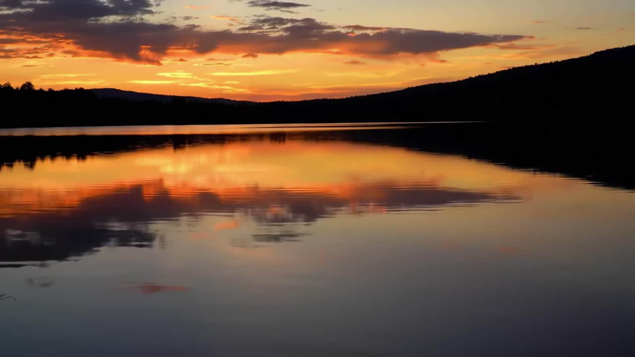 A stunning sunset casts vibrant colors over the lake, creating mesmerizing reflections on the water. Mountains silhouette against the colorful sky as day transitions to night.