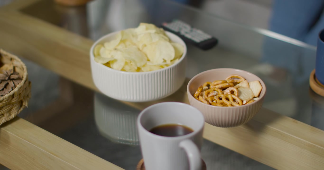 Die Kamera zieht sich zurück und steigt auf, enthüllt eine gerippte Tasse auf einem Untersetzer, Chips, Snacks und eine Fernbedienung, die das Arrangement zeigen.