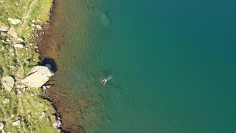 Top-down-drone-footage-of-a-man-swimming-near-the-shore-of-a-lake-in-the-mountains