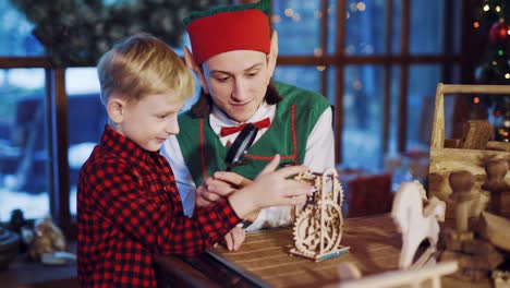 an elf with long ears and a red hat is holding a magnifying glass in his hand and showing a little boy a funny toy on the background of festive decorations and a christmas tree near the window in the residence of santa claus