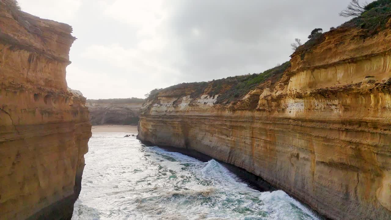 Aerial footage of rugged cliffs and ocean waves at Loch Ard Gorge, Port Campbell, Australia. Overcast lighting enhances the dramatic landscape