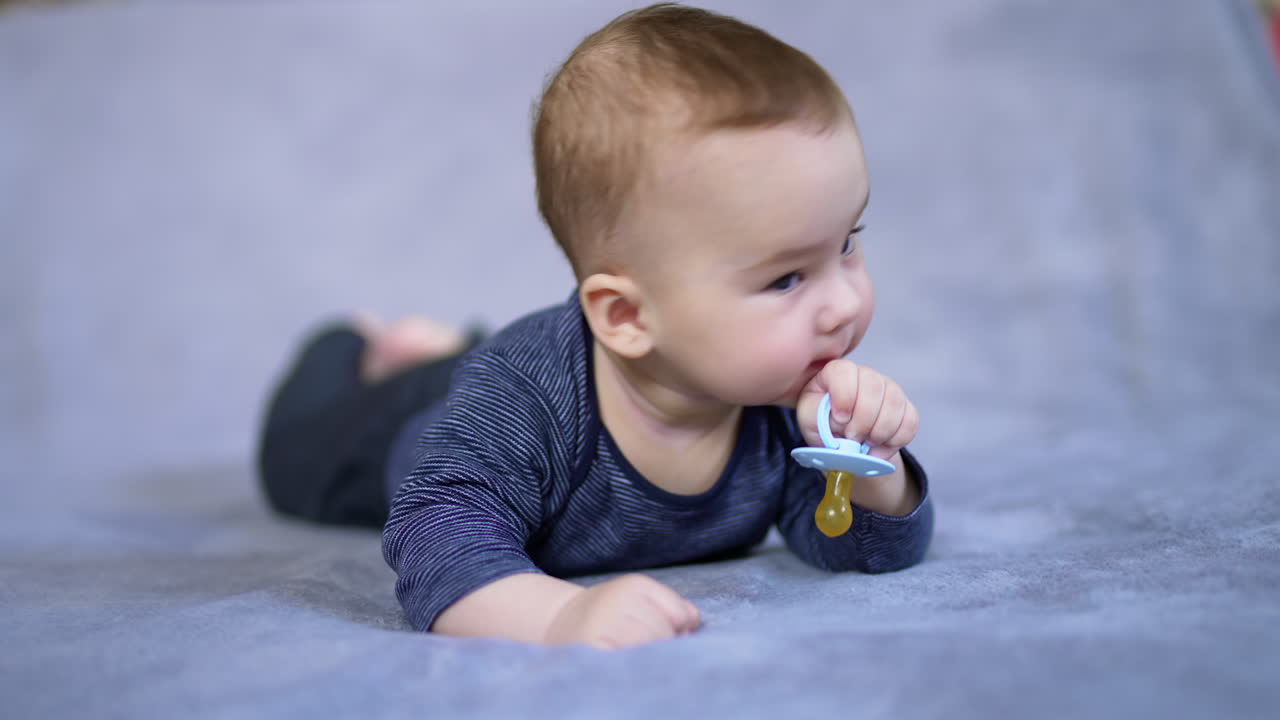 Sweet tiny boy lies on bed holding a pacifier in his hand. Beautiful kid turns his head to look around and smiles adorably. Close up.