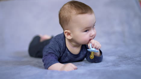 sweet tiny boy lies on bed holding a pacifier in his hand. beautiful kid turns his head to look around and smiles adorably. close up.