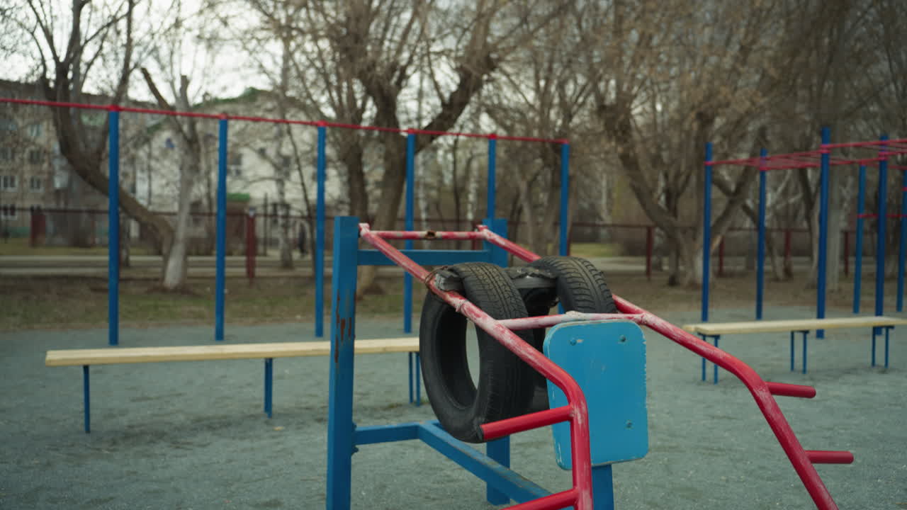 재활용 된 타이어와 금속 막대기로 만들어진 파란색과 빨간색 운동 장비를 볼 수 있는 야외 체육관 설정의 클로즈업, 빈 나무와 인근 벤치