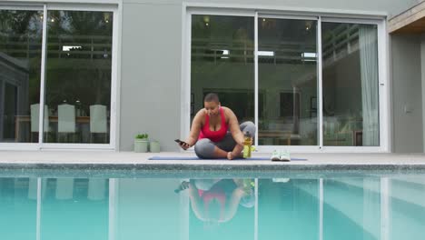 happy african american plus size woman practicing yoga, using smartphone next to swimming pool