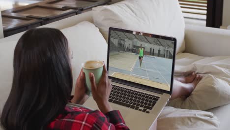 Composite-of-woman-sitting-at-home-holding-coffee-watching-athletics-long-jump-event-on-laptop