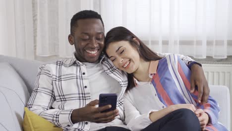 happy couple african man and caucasian woman sitting together on sofa at home and laughing while looking at phone.