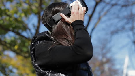 mujer joven con abrigo negro cálido con gafas de realidad virtual en el parque de otoño temprano