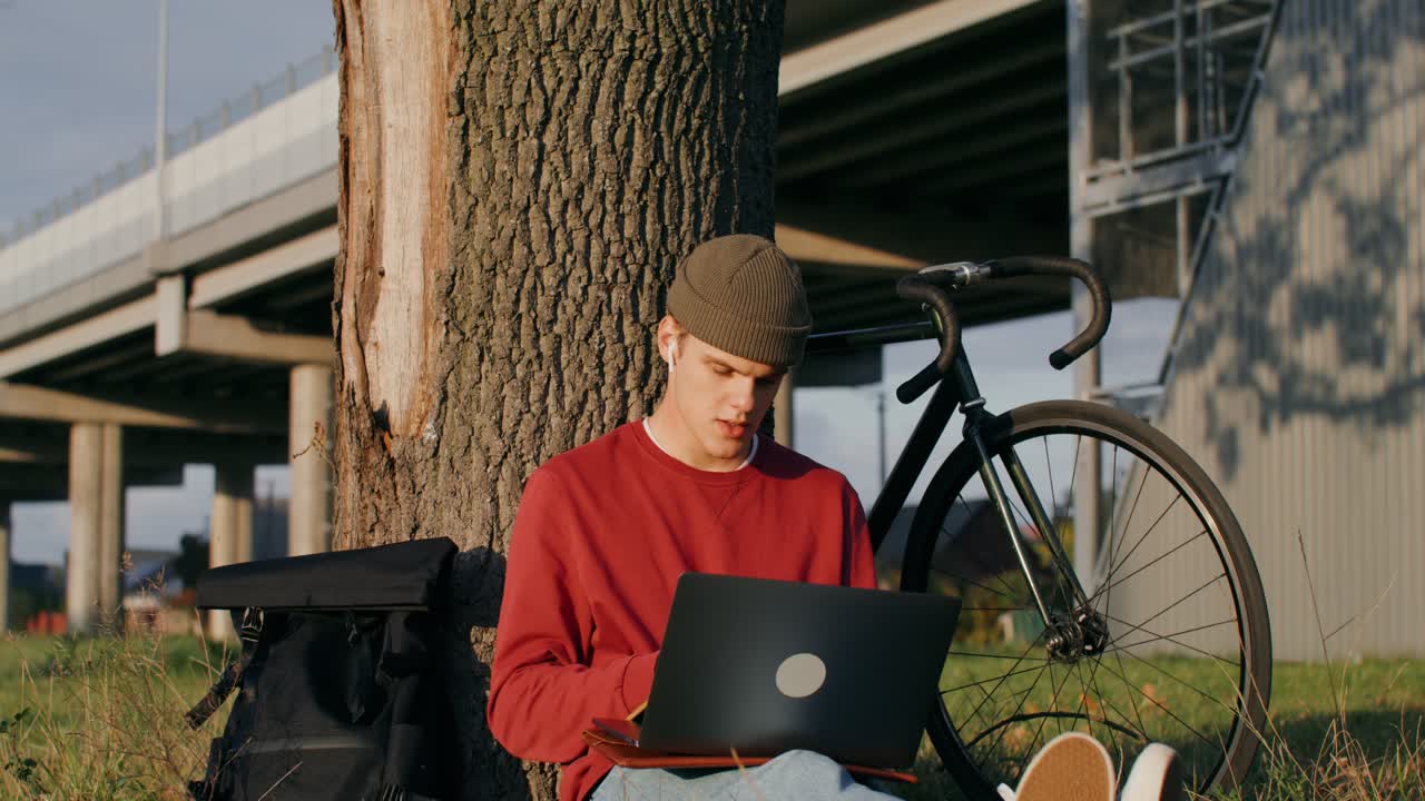 hombre trabajando en una computadora portátil al aire libre bajo un árbol por una bicicleta