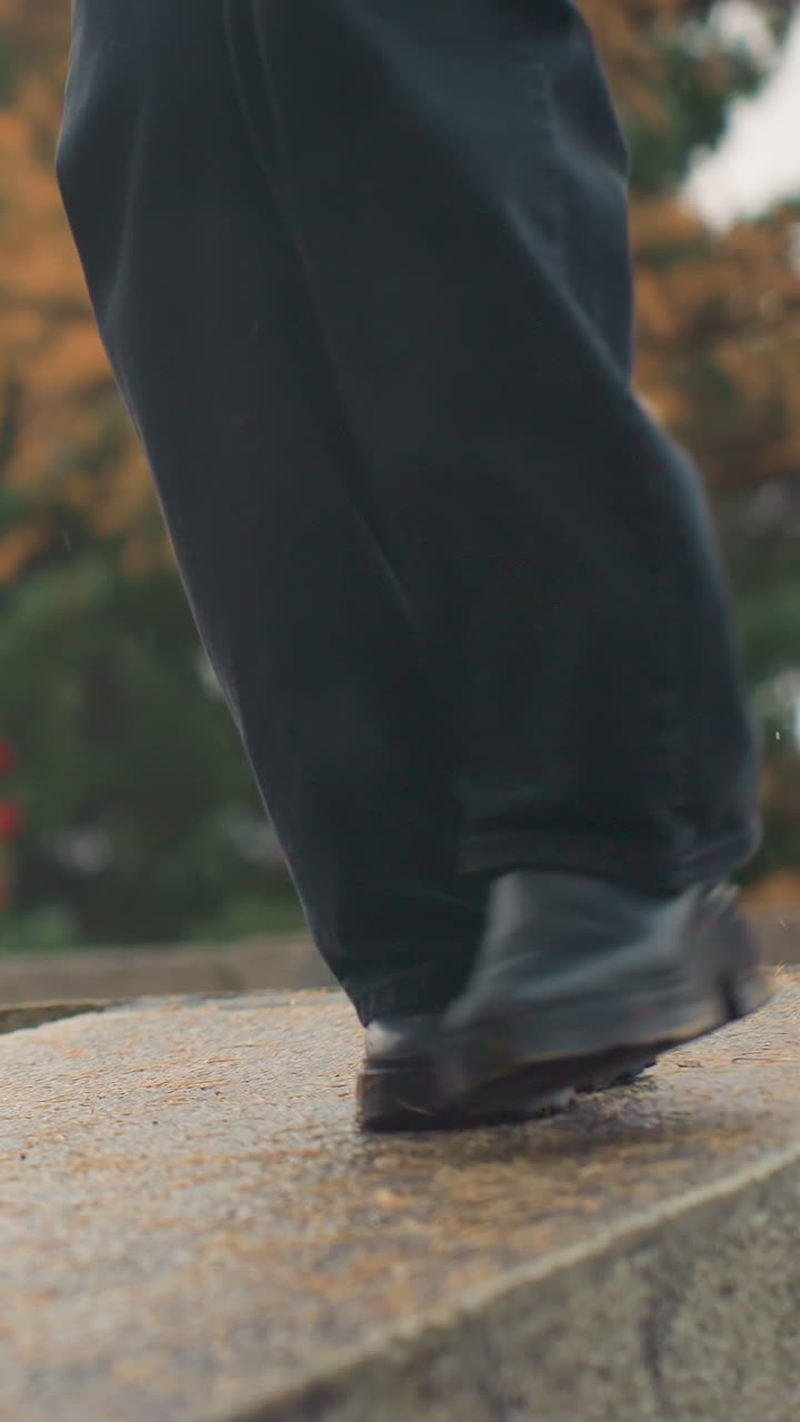 Close-up view of person in black trousers and black boots carefully walking on wide wet stone path during rainy autumn day, maintaining balance amidst the rain and leaves
