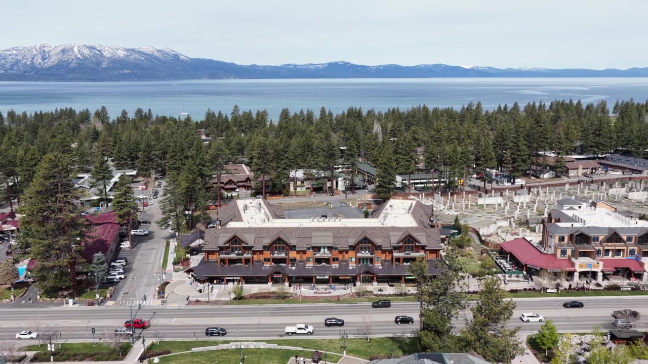 Cittadina alpina di South Lake Tahoe con edifici in stile lodge rustico lungo il viale principale, fitta foresta di pini e il luccicante Lago Tahoe con cime innevate sullo sfondo. California, USA. Ampia ripresa aerea laterale