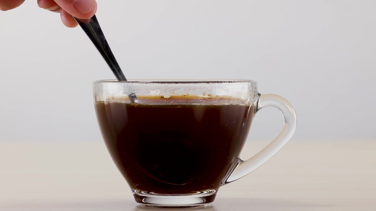 Primer plano de una mano revolviendo café negro caliente con una cuchara en una taza de cristal.