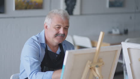 a female teacher shows a retired man how to draw a picture with paints and a brush at courses for the elderly. a senior man draws a picture to a group of pensioners