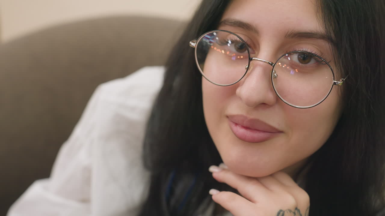 Close up of fair skin lady with round glasses resting chin on hand, smiling softly while looking down, delicate reflection of light visible on lens, creating calm and charming atmosphere