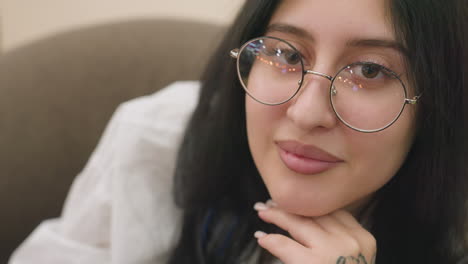 close up of fair skin lady with round glasses resting chin on hand, smiling softly while looking down, delicate reflection of light visible on lens, creating calm and charming atmosphere