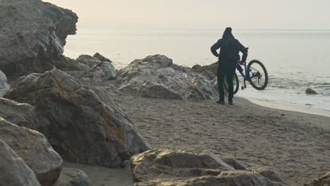 man-walks-his-bicycle-along-a-sandy-and-rocky-beach,-he-carefully-carries-his-bike-over-rocks,-showcasing-his-determination-and-resilience