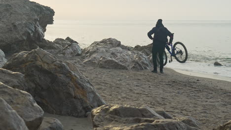 man walks his bicycle along a sandy and rocky beach, he carefully carries his bike over rocks, showcasing his determination and resilience