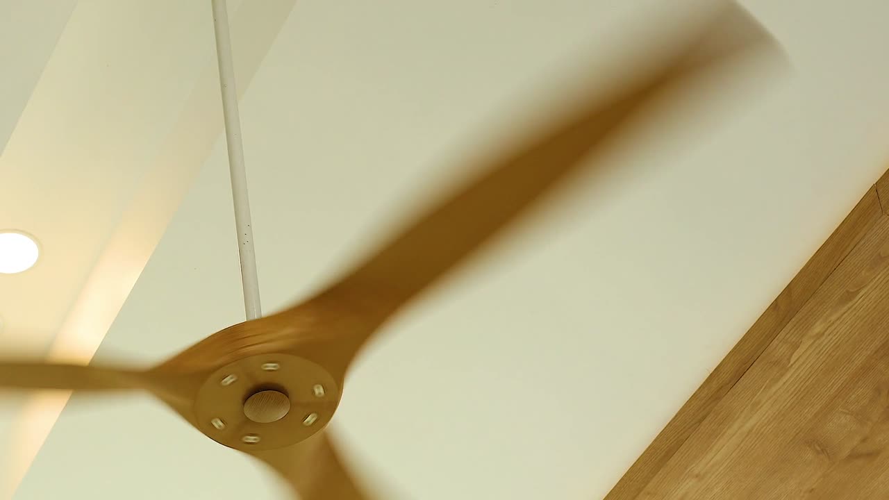 A wooden ceiling fan rotates steadily, casting shadows on a white ceiling with recessed lighting.
