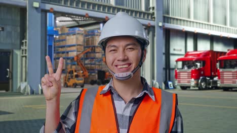 close up of asian male engineer with safety helmet smiling and showing peace gesture while standing , outside of logistics distributions warehouse