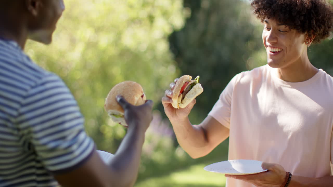 Eating burgers, two multiracial male friends enjoying outdoor hangout, smiling and having fun
