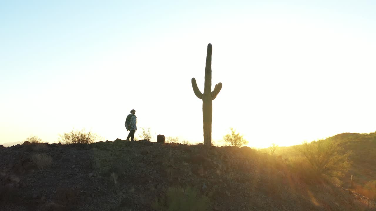 un forajido del salvaje oeste con un poncho camina a lo largo de una cresta del desierto pasando un cacto saguaro