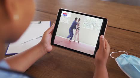 Composite-of-woman-sitting-at-table-with-face-mask,-watching-judo-match-on-tablet