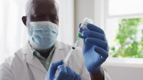 male african american doctor wearing face mask pulling out covid-19 vaccine into syringe