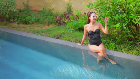 asian woman sitting on swimming pool edge with black swimsuit and vintage sunglasses and legs crossed