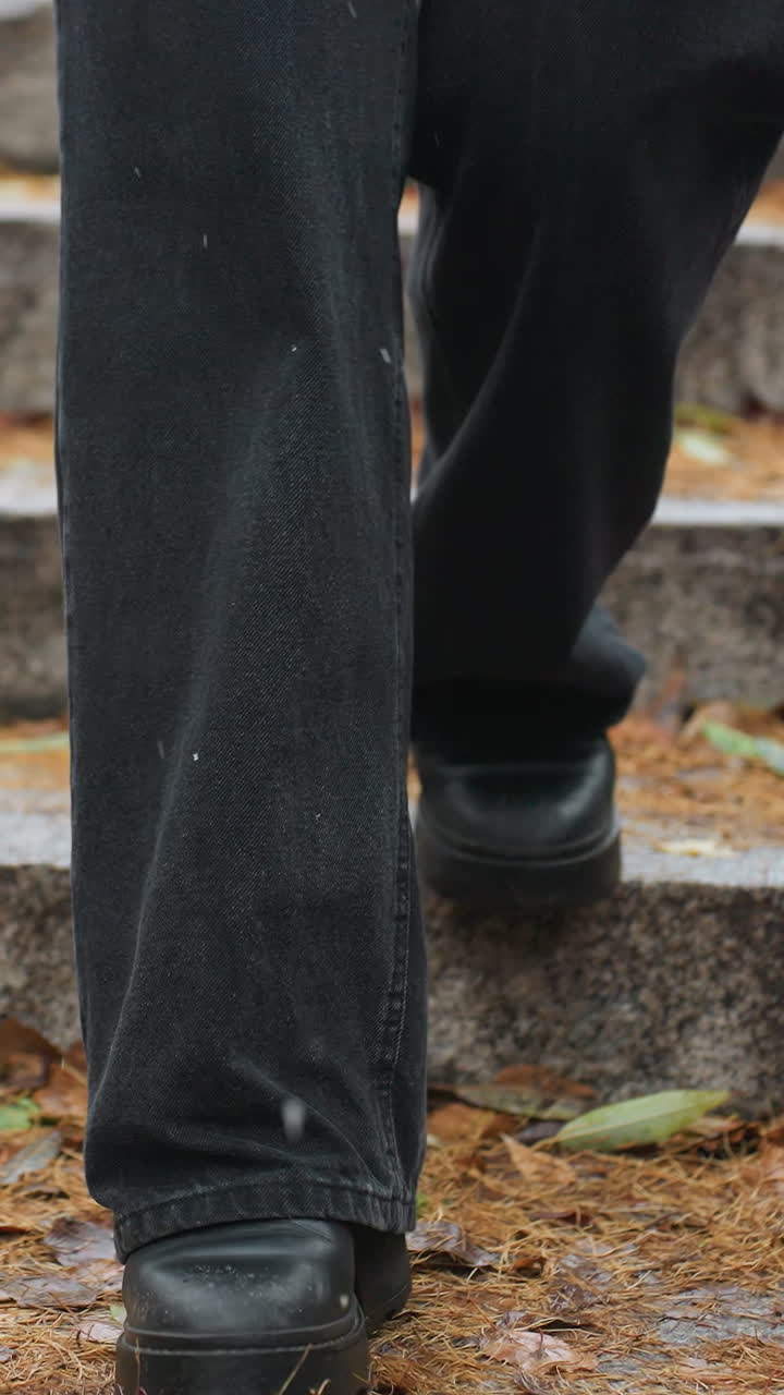 Person's legs walking down stone steps during light snowfall, wearing black jeans and black boots, with colorful autumn leaves scattered along the path, creating a seasonal vibe