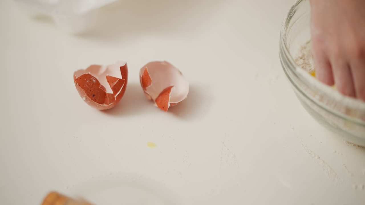close up of cracked brown egg shell lying on white surface near glass bowl containing flour and raw egg, slight egg smear visible on table, capturing hands-on baking preparation detail in kitchen
