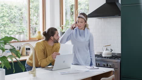 Diverse-couple-using-laptop-and-smartphone-in-kitchen-at-home,-in-slow-motion