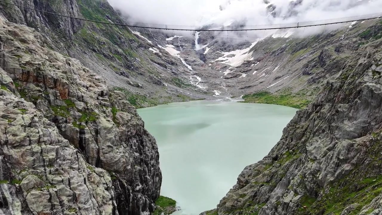 Catturando le acque del lago glaciale e del ponte di Trift, con l'acqua che scende a cascata, incastonato tra aspre scogliere nelle Alpi svizzere innevate, Triftbrücke Triftgletscher, Berna, Svizzera, ripresa con drone