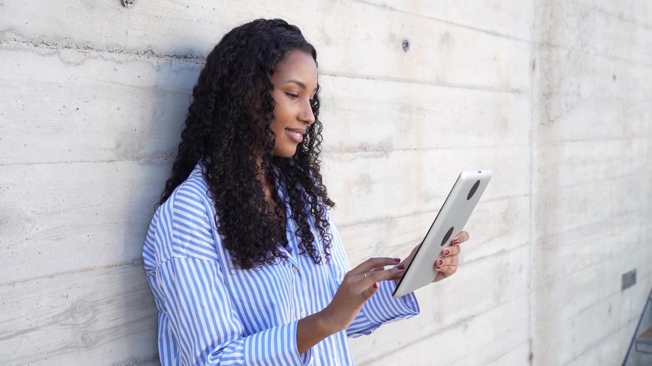 A young woman is actively engaged with her tablet device while positioned against a concrete wall