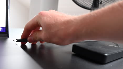 a laptop user plugs an external hardrive with a black cable to his computer on his desk