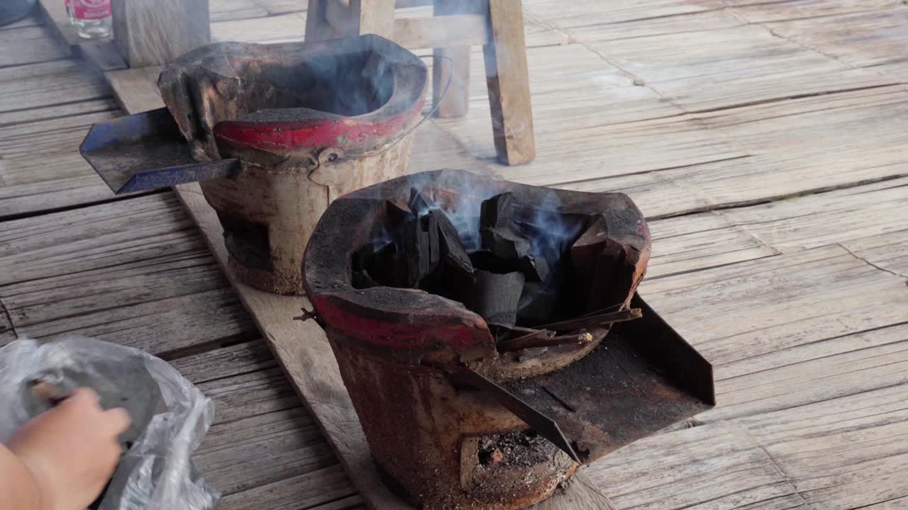 Close-up of a person refilling charcoal in a traditional Thai clay charcoal stove with embers smoking for barbecue cooking. The scene captures the essence of rural Thai cooking methods