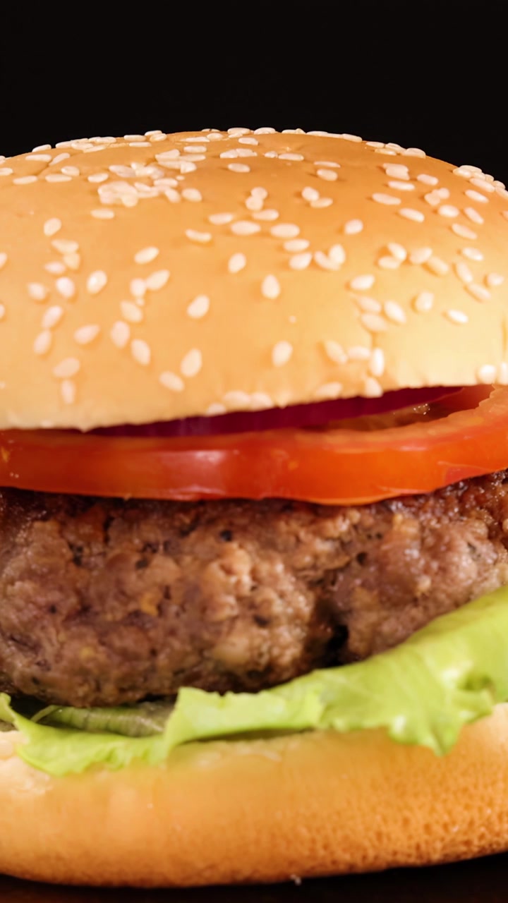 Una mano posa delicatamente un panino ai semi di sesamo sopra un hamburger di manzo con lattuga, pomodoro e cipolla, sotto una brillante illuminazione da studio su uno sfondo nero.
