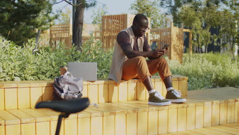 young black man sitting in park and using smartphone