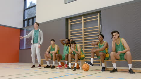 multiracial male basketball players with coach in gym, supporting and sitting on bench
