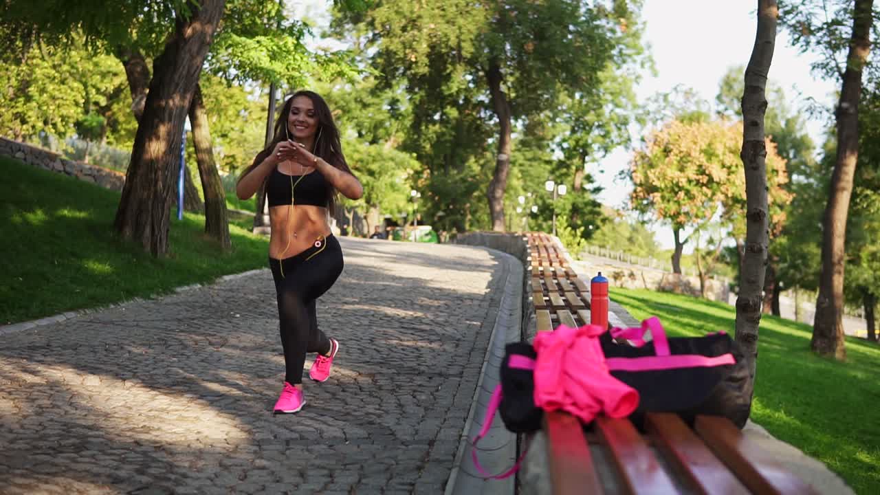 chica de fitness está haciendo estocadas en el parque en verano escuchando la música en los auriculares. entrenamiento en un día soleado en cámara lenta