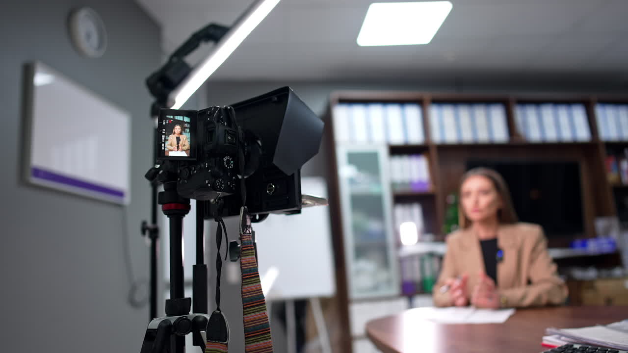 Professional camera on tripod filming a young woman in beige jacket sitting at desk. Influencer creating video for a blog. Blurred backdrop.