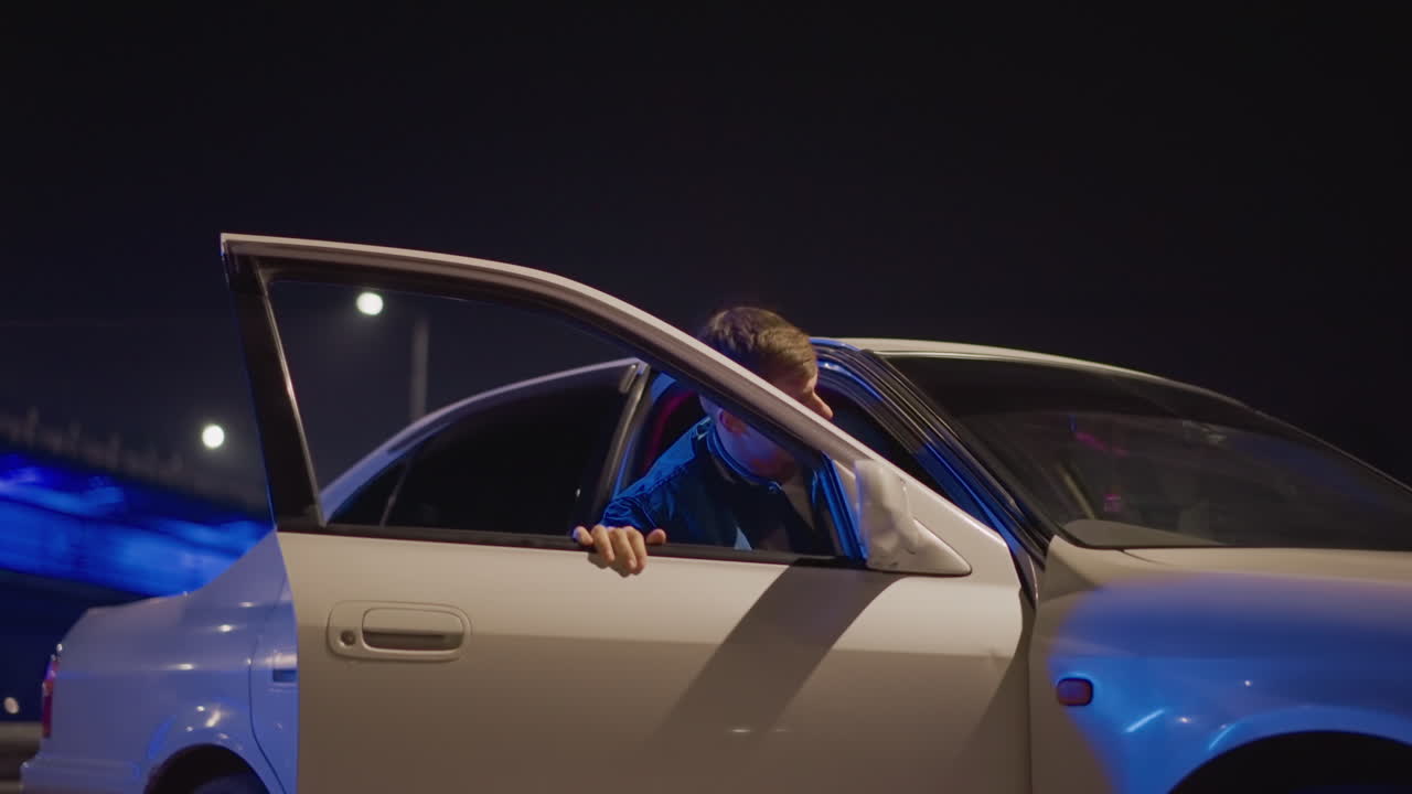 man approaches parked car under bridge lit by cool blue LEDs, white sedan waiting by guardrail, jacketed figure walks up, opens door and leans toward interior, brief tense exchange and cautious