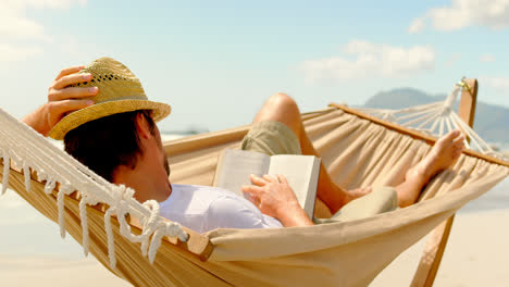 Side-view-of-caucasian-man-reading-in-hammock-at-beach-4k